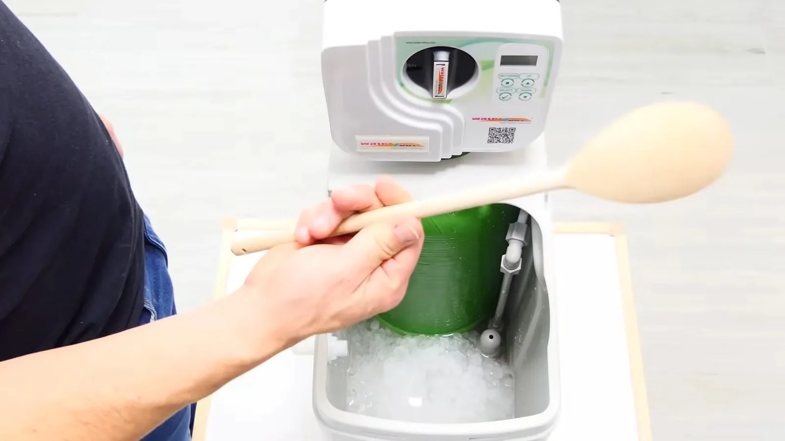 A hand holding a wooden spoon over a water softener's open brine tank, where a crust of salt sits around a green cylinder.