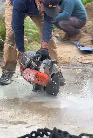 A man in a cap cutting a concrete slab with an orange saw while another crouches in the background.
